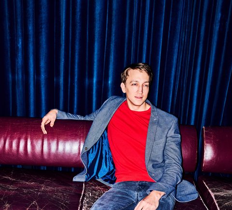 Ein Mann in blauem Blazer und rotem T-Shirt sitzt entspannt auf einem roten Sofa mit einem Drink auf dem Tisch.