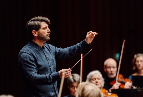 Ein Dirigent führt ein Orchester in einem Konzertsaal. Er hebt den Taktstock, während die Musiker aufmerksam sind.