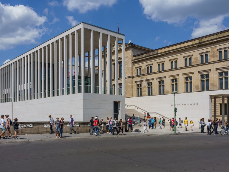 Das Foto zeigt ein modernes Gebäude mit Säulen und ein klassisches Gebäude. Menschen stehen davor unter einem blauen Himmel.