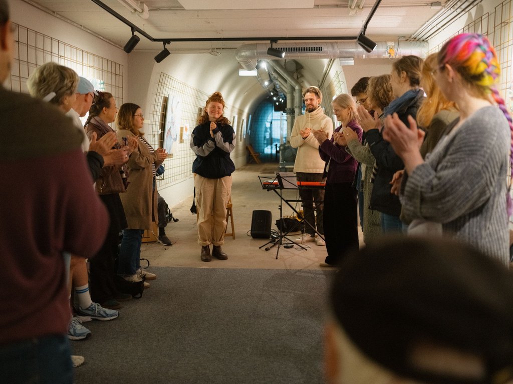 Eine Gruppe von Menschen steht in einem langen, beleuchteten Korridor und applaudiert. Ein Musiker spielt. Die Atmosphäre ist lebhaft.