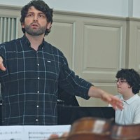 Ein Dirigent leitet eine Probe in einem Musikraum. Ein Pianist spielt im Hintergrund. Partituren sind im Vordergrund.