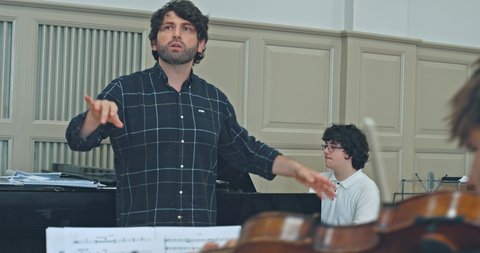 Ein Dirigent leitet eine Probe in einem Musikraum. Ein Pianist spielt im Hintergrund. Partituren sind im Vordergrund.  © Jasper Techel