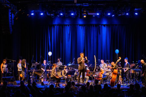 Ein Orchester auf einer Bühne, beleuchtet von blauem Licht. Ein Dirigent steht in der Mitte, Musiker spielen verschiedene Instrumente.