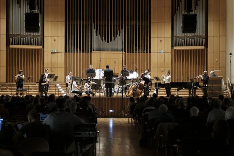 Das Kammerorchester Alarm Will Sound steht auf der Bühne der Aula in der Universität Bonn. Der Dirigent steht in der Mitte, während das Publikum aufmerksam zuhört.