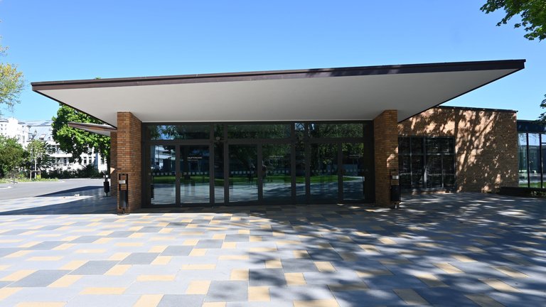 Modernes Gebäude mit Glasfassade und weit überstehendem Dach. Es steht auf einem schachbrettartig gepflasterten Platz unter blauem Himmel.