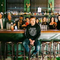 Eine Gruppe von Menschen, das Vokalensemble Roomful of Teeth, steht hinter einer Bar mit verschiedenen Flaschen im Hintergrund. Eine Person, Gabriel Kahane, sitzt auf einem Stuhl vorne.