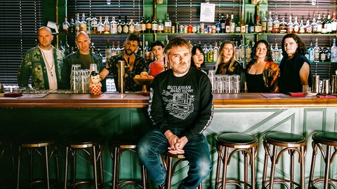 Eine Gruppe von Menschen, das Vokalensemble Roomful of Teeth, steht hinter einer Bar mit verschiedenen Flaschen im Hintergrund. Eine Person, Gabriel Kahane, sitzt auf einem Stuhl vorne.  © Charlie Weinmann