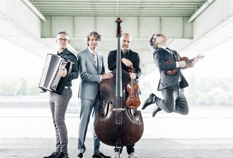 Vier Musiker in Anzügen posieren unter einer Brücke mit Instrumenten: Akkordeon, Kontrabass, Violine und einer springt mit Viola.
