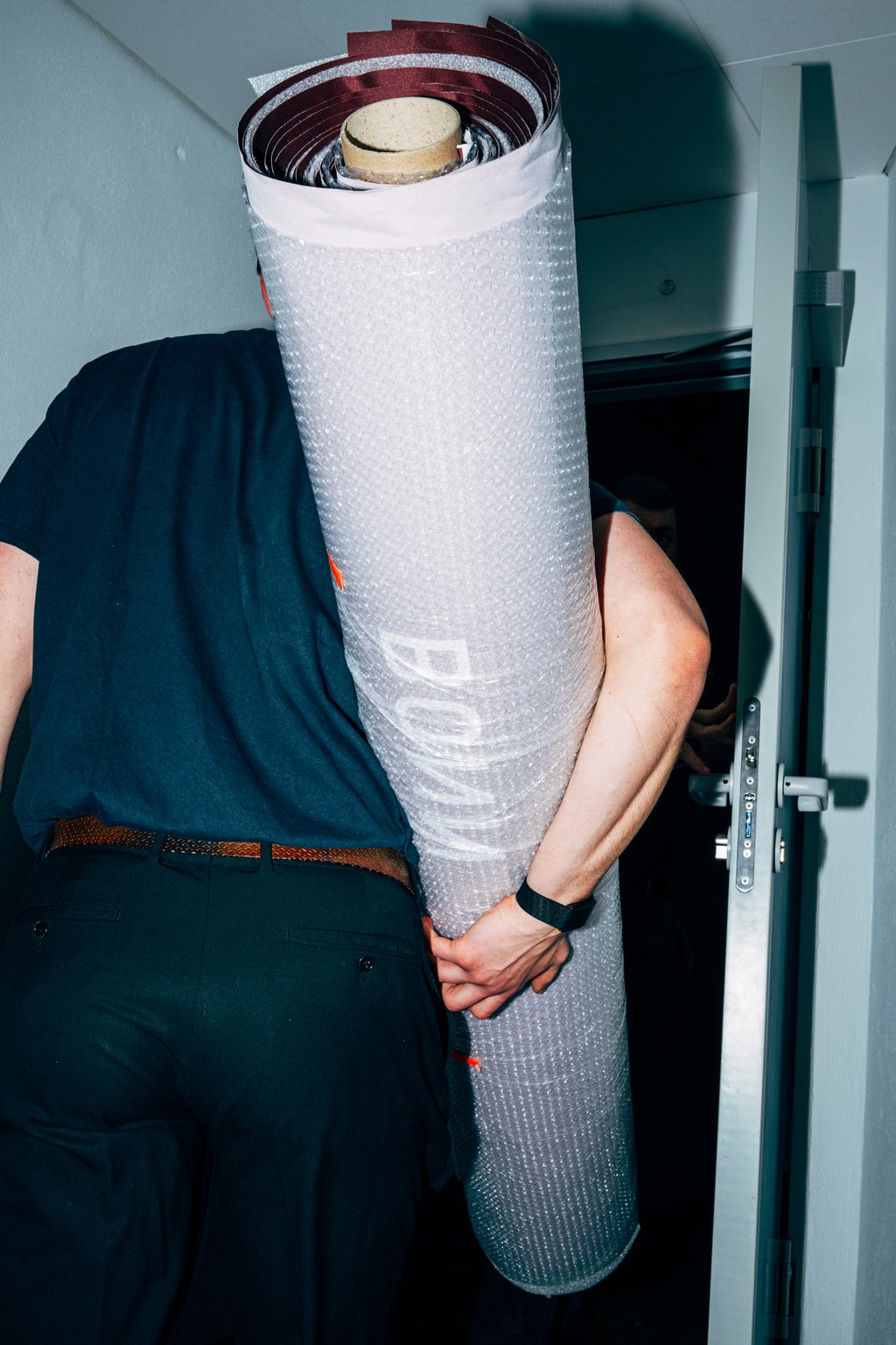 Eine Person trägt eine zusammengerollte große Leinwand durch eine Tür in ein beleuchtetes Zimmer.