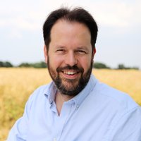 Ein Mann steht tagsüber vor einem Feld. Er trägt ein helles Hemd. Er blickt in die Kamera und lächelt.