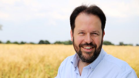 Ein Mann steht tagsüber vor einem Feld. Er trägt ein helles Hemd. Er blickt in die Kamera und lächelt.  © Marshall Light Studio