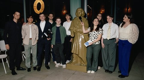 Ein Gruppenbild von jungen Leute, die in einer Reihe um einer große Beethovenstatue herum stehen. Im Hintergrund eine Glasfront mit schwarzem Nachthimmel. Eine junge Frau hält Beethoven ein Mikrofon vor das Gesicht.