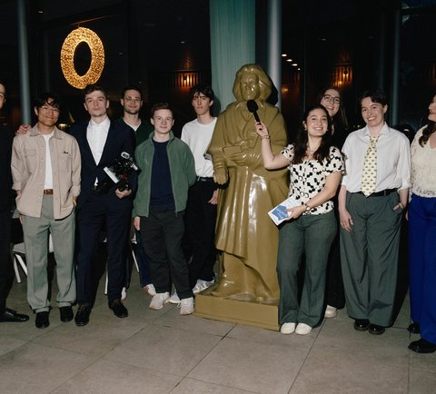 Ein Gruppenbild von jungen Leute, die in einer Reihe um einer große Beethovenstatue herum stehen. Im Hintergrund eine Glasfront mit schwarzem Nachthimmel. Eine junge Frau hält Beethoven ein Mikrofon vor das Gesicht.