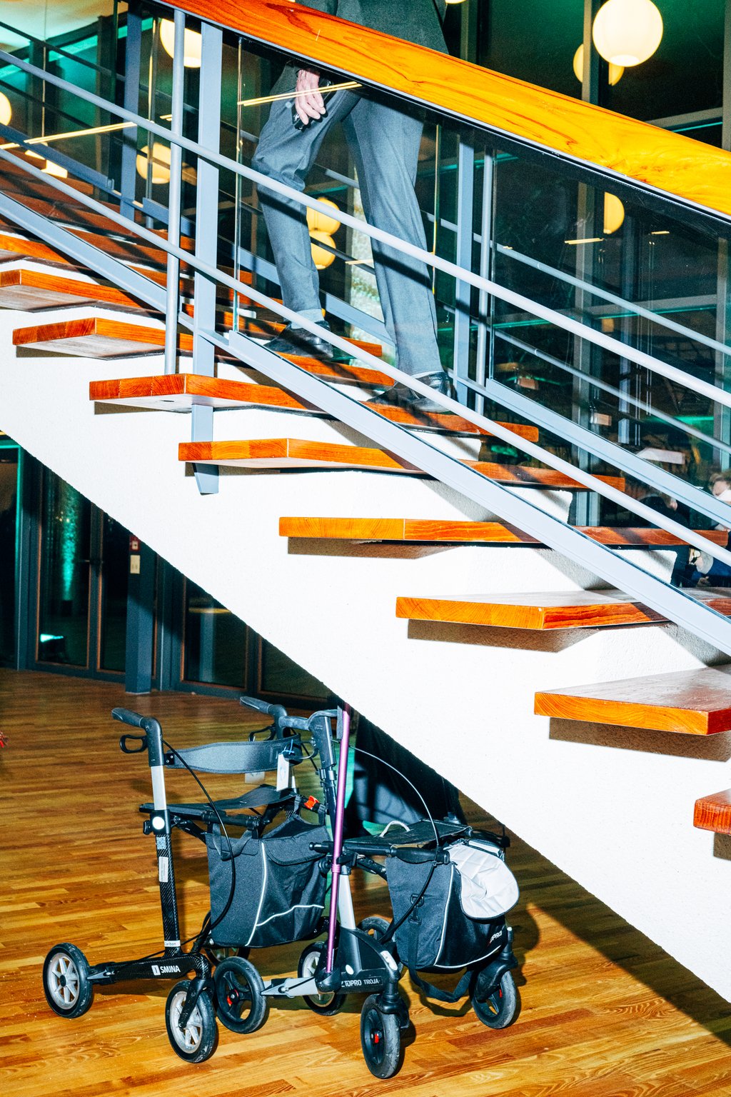 Ein Rollator steht unten an einer modernen Holztreppe, während eine Person darauf hinaufgeht. Reflektierende Oberflächen im Hintergrund.