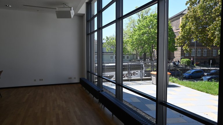 Moderner Raum mit großer Fensterfront, die Blick auf eine sonnige Straße mit Bäumen und einem Gebäude bietet.