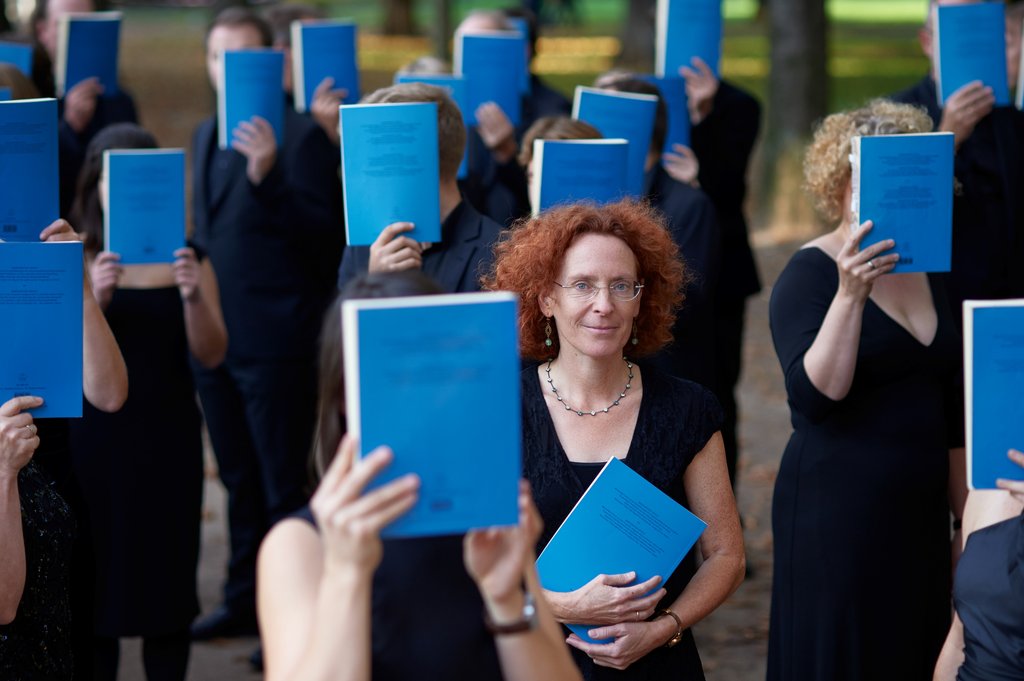 Eine Gruppe von Menschen in schwarzer Kleidung hält blaue Chorbücher hoch. Eine Frau steht lächelnd in der Mitte.