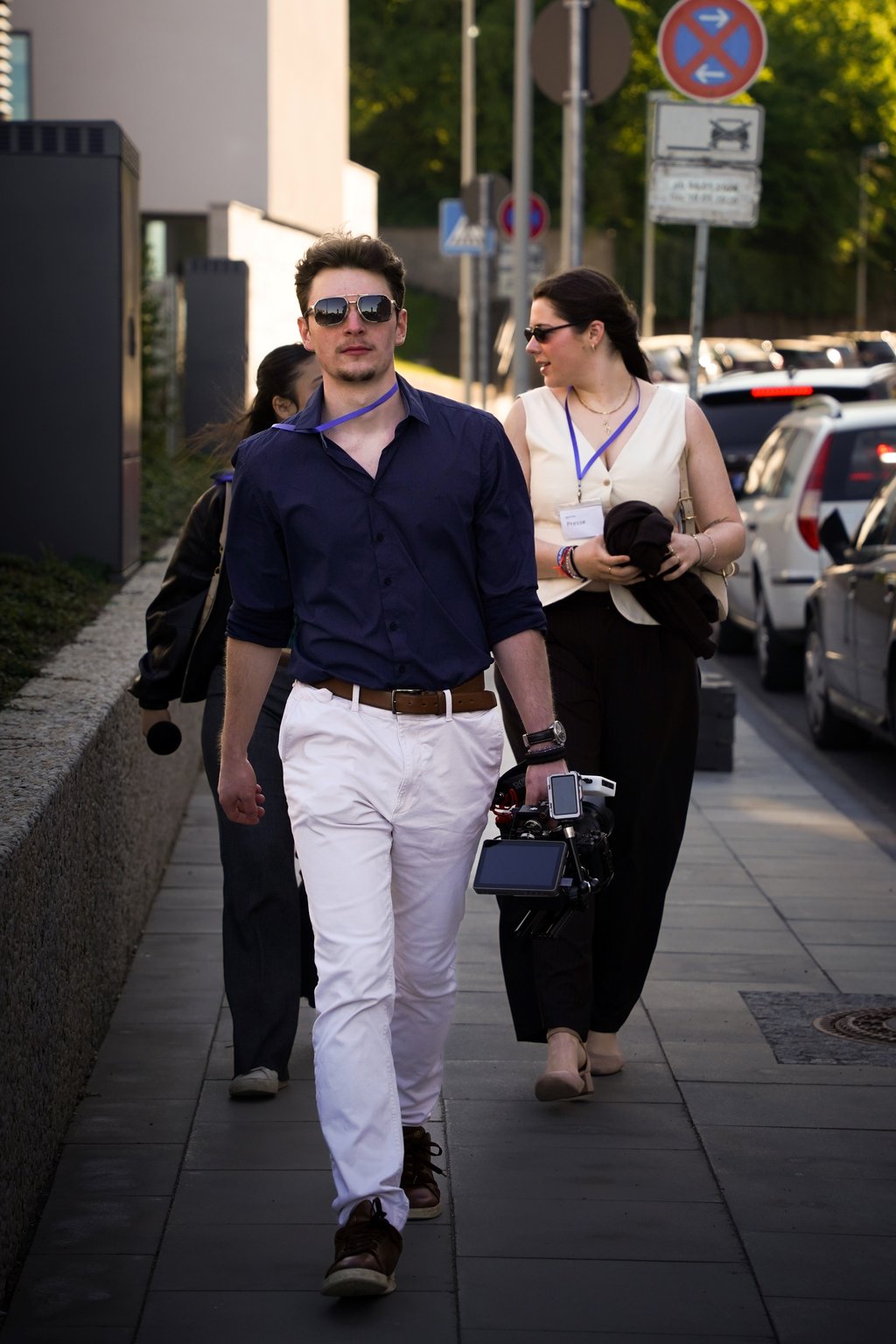 Mann in blauer Kleidung und weißer Hose mit Kamera in der Hand, geht auf Gehweg mit anderen Personen im Hintergrund.