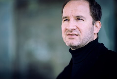 Ein Mann im Porträt, er schaut ernst an der Kamera vorbei. Er trägt einen schwarzen Rollkragenpullover. Der Hintergrund ist türkis-grau.
