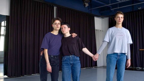 Drei Personen stehen eng zusammen in einem Tanzstudio, halten sich an den Händen und Schultern, lässig in Jeans und T-Shirts gekleidet.