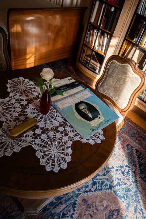 Ein runder Holztisch mit einem weißen Spitzendeckchen, Schallplatten mit einem Porträt und einer Einzelrose in einer Vase.