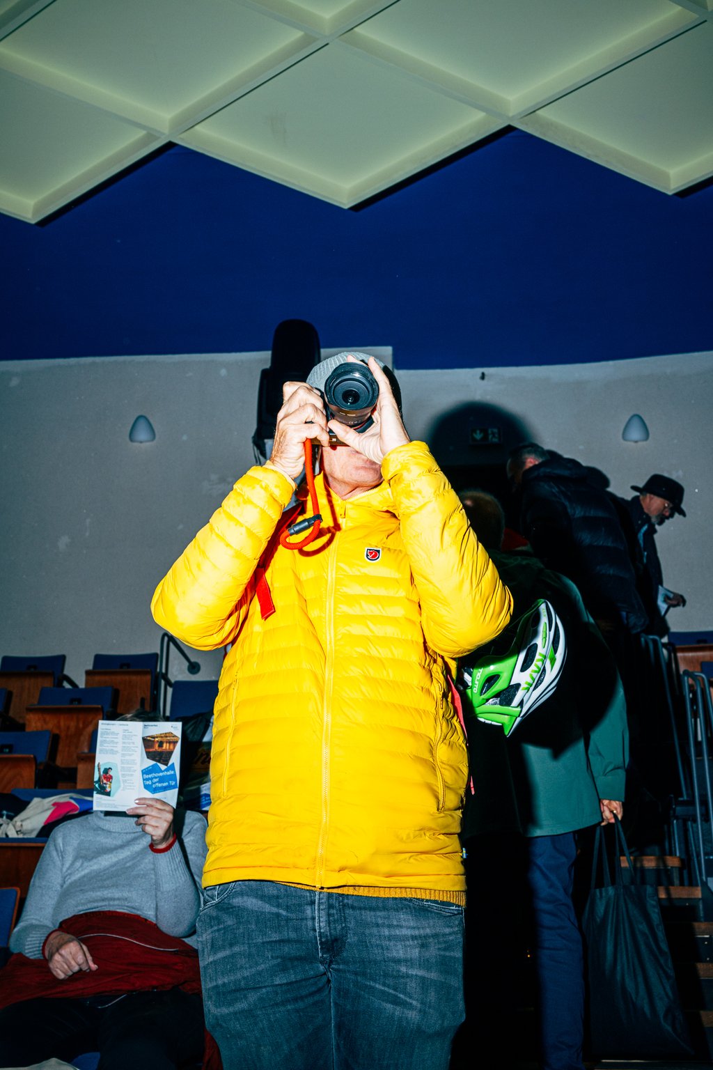 Eine Person in einer gelben Jacke macht ein Foto in einem Veranstaltungsraum. Andere Menschen sitzen oder bewegen sich im Hintergrund.