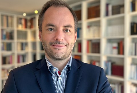 Ein Mann in einem Anzug steht vor einem Bücherregal. Das Regal ist mit Büchern gefüllt und der Raum ist gut beleuchtet.