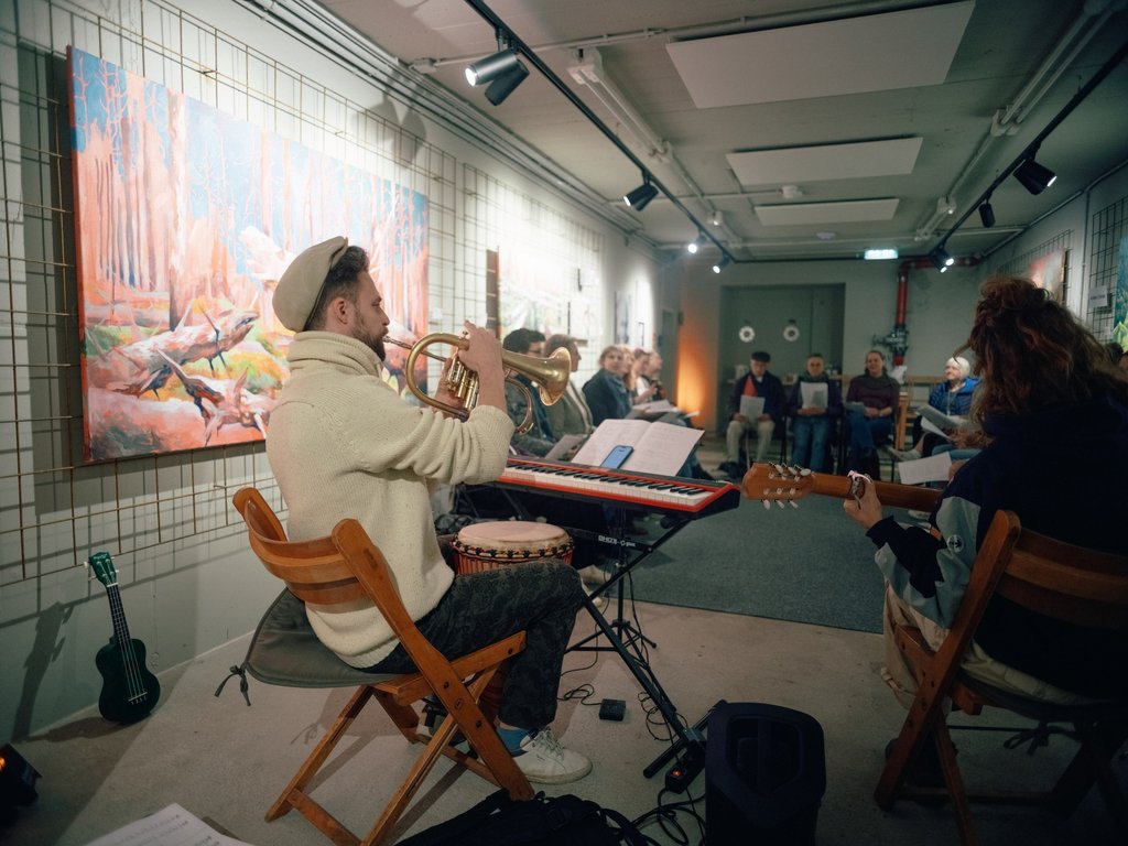 Ein Chor singt in einer Galerie. Gemälde hängen an den Wänden. Die Atmosphäre wirkt entspannt und künstlerisch.