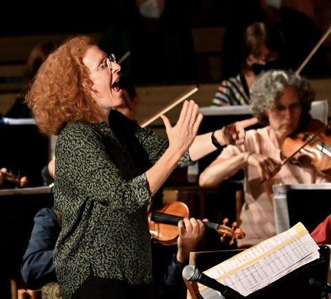 Eine Dirigentin mit lockigem Haar leitet ein Orchester. Mehrere Musiker spielen Streicherinstrumente, konzentriert auf die Musik.