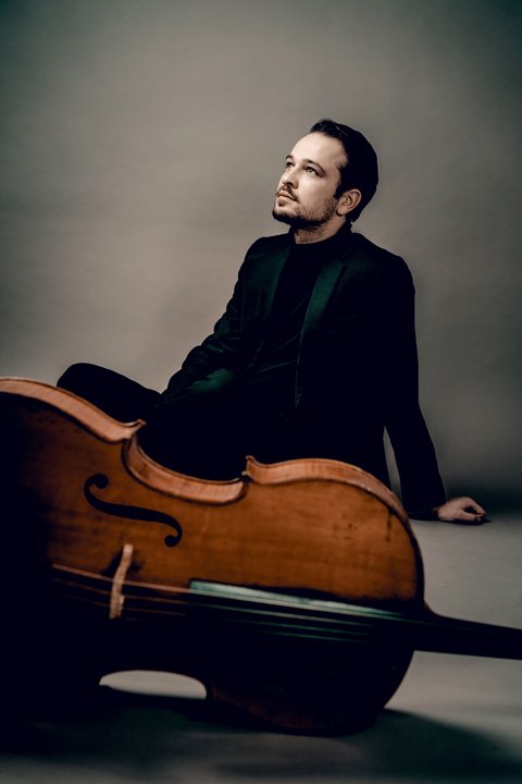 Ein Mann sitzt entspannt und schaut nachdenklich, während er neben einem großen Cello sitzt, in einem professionellen Studio.  © Andrej Grilc