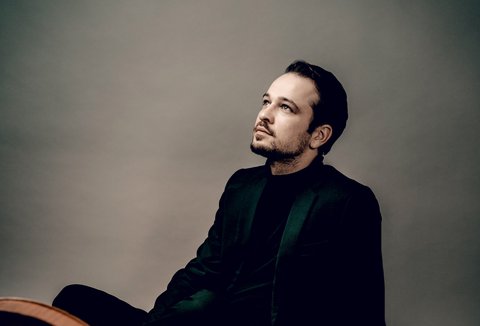 Ein Mann sitzt entspannt und schaut nachdenklich, während er neben einem großen Cello sitzt, in einem professionellen Studio.