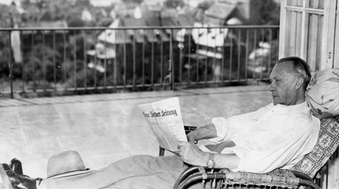 Eine Person sitzt auf einem Balkon und liest eine Zeitung. Im Hintergrund ist eine Landschaft mit Gebäuden zu sehen.
