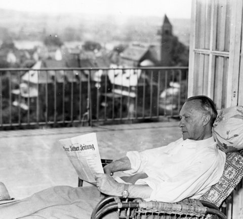 Eine Person sitzt auf einem Balkon und liest eine Zeitung. Im Hintergrund ist eine Landschaft mit Gebäuden zu sehen.