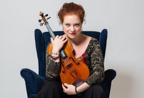 Eine Frau mit einer Viola sitzt auf einem Sessel. Sie trägt ein gemustertes Oberteil und blickt in die Kamera.