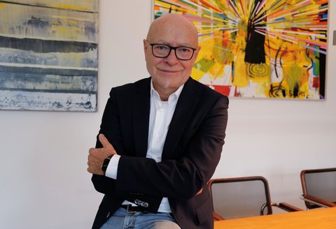 Ein Mann steht vor mehreren Kunstwerken. Er trägt einen Anzug. Seine Arme sind verschränkt. Er blickt in die Kamera und lächelt.