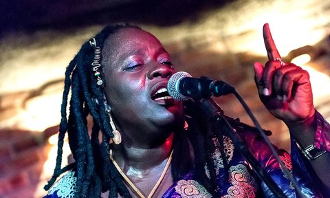 Eine Frau mit langen Dreadlocks singt leidenschaftlich in ein Mikrofon auf der Bühne. Der Hintergrund ist stimmungsvoll beleuchtet.  © Kora Music Festival