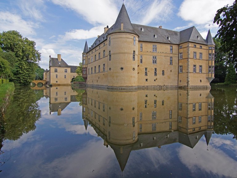 Ein historisches Schloss mit Türmen spiegelt sich in einem ruhigen Wassergraben. Der Himmel ist klar und blau.