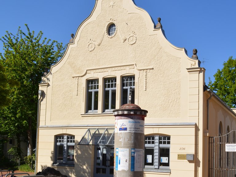 Historisches Gebäude mit einer dekorativen Fassade und einer Werbesäule im Vordergrund. Der Himmel ist klar und blau.