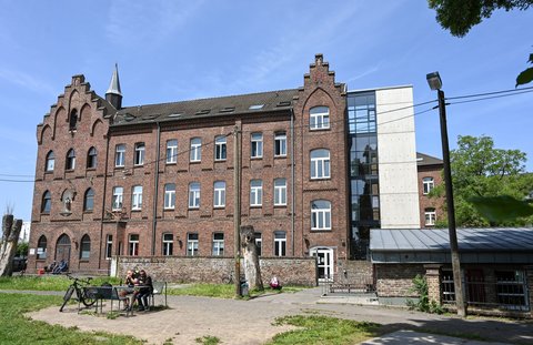 Historisches Backsteingebäude mit modernen Glas- und Betonelementen. Vor dem Gebäude sitzen Menschen im Freien auf einem Platz mit Grünflächen.