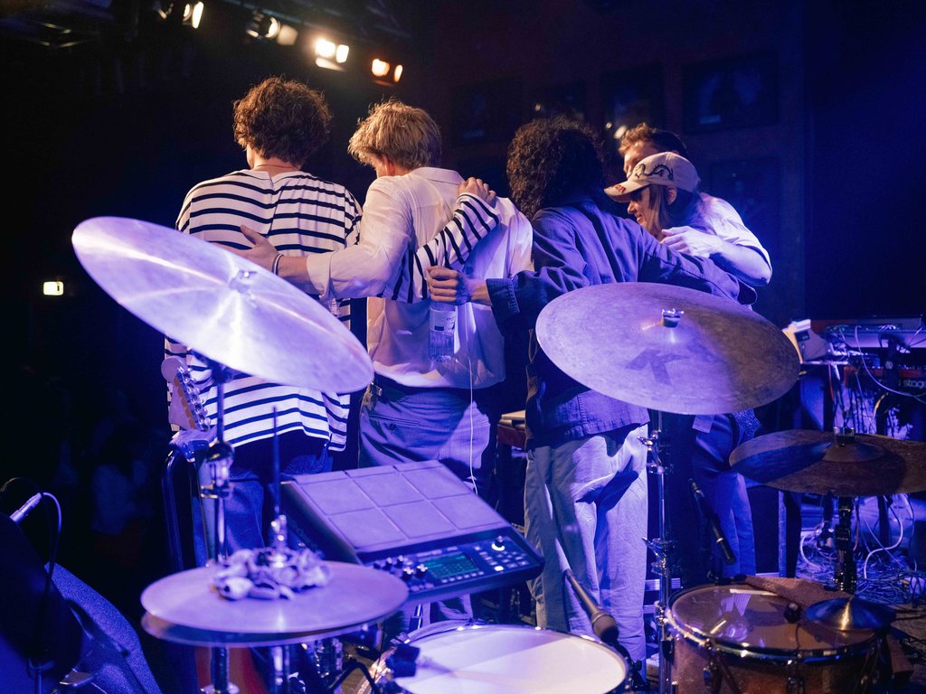 Musikergruppe auf der Bühne, von hinten fotografiert, umarmt sich nach einem Auftritt. Schlagzeug und Lichter im Vordergrund sichtbar.