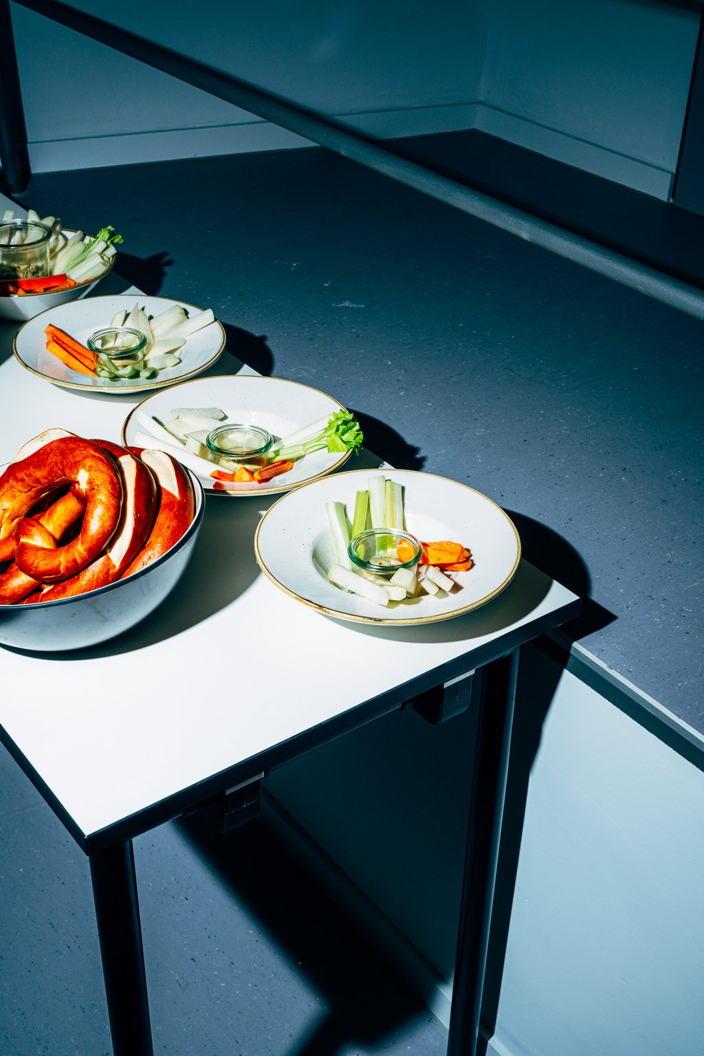 Ein gedeckter Tisch mit tellern Salaten, Brezeln und Gemüse, darunter Sellerie und Karotten, in heller Beleuchtung.