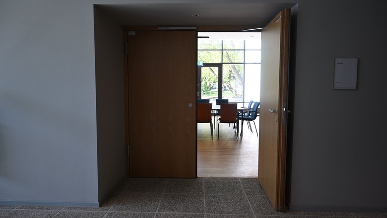 Eine geöffnete Tür führt in einen Konferenzraum mit Stühlen und Tischen. Licht fällt durch die Fenster im Hintergrund.