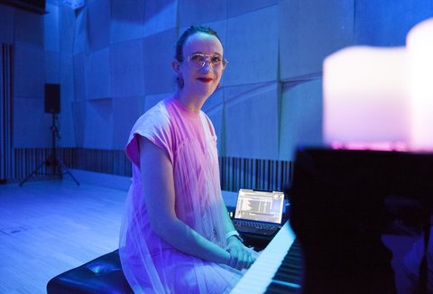 Eine Person in rosa Kleidung spielt Klavier in einem blauen, stimmungsvoll beleuchteten Raum mit Kerzen im Vordergrund.