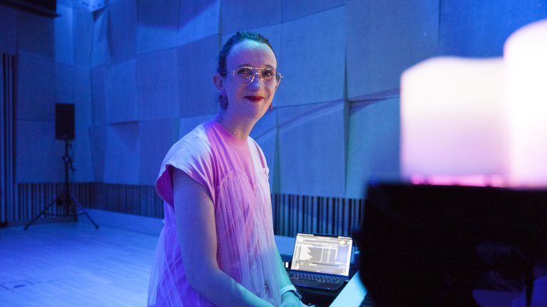 Eine Person in rosa Kleidung spielt Klavier in einem blauen, stimmungsvoll beleuchteten Raum mit Kerzen im Vordergrund.