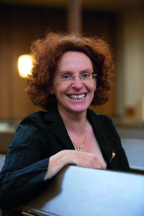 Eine Frau mit lockigem, rotem Haar lächelt und trägt ein dunkles Oberteil. Sie sitzt in einem beleuchteten Raum.  © Ralf Bauer
