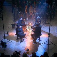 Ein Streichquartett spielt in einem dunklen Raum, umgeben von sitzendem Publikum. Stimmungsvolle Lichter beleuchten die Musiker von oben.