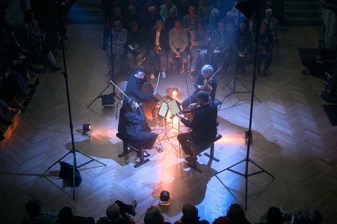 Ein Streichquartett spielt in einem dunklen Raum, umgeben von sitzendem Publikum. Stimmungsvolle Lichter beleuchten die Musiker von oben.