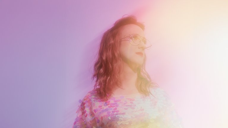 Ein unscharfes Porträt einer Person mit langen Haaren und Brille, in regenbogenfarbenem Licht gehüllt, vor lila Hintergrund.