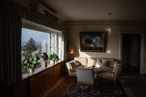 Gemütliches Wohnzimmer mit Sesseln, bestückten Bücherregalen und Gemälde an der Wand. Durch das große Fenster strömt Tageslicht.