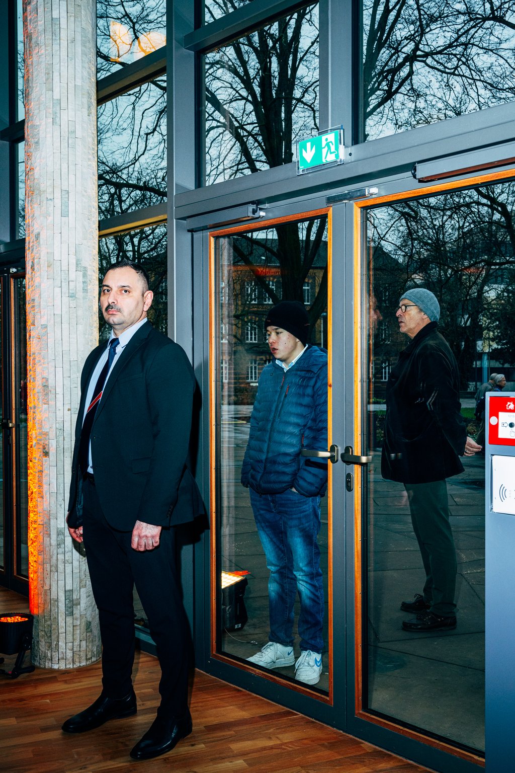 Ein Wachmann steht vor einer Glastür, während zwei Personen draußen stehen, beide in Winterkleidung. Die Szene ist beleuchtet.