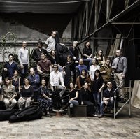 Eine große Gruppe von Menschen posiert gemeinsam in einem industriellen Raum. Einige halten Musikinstrumente, andere stehen lässig.